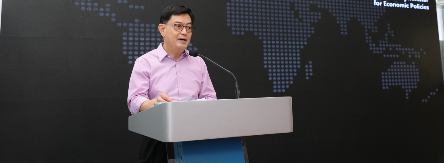 Man in glasses and pink shirt stands at podium with world map background.